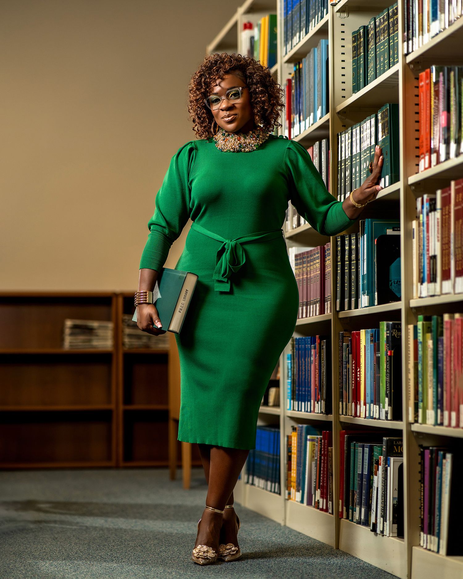 Person in green dress poses with book in library between bookshelves.