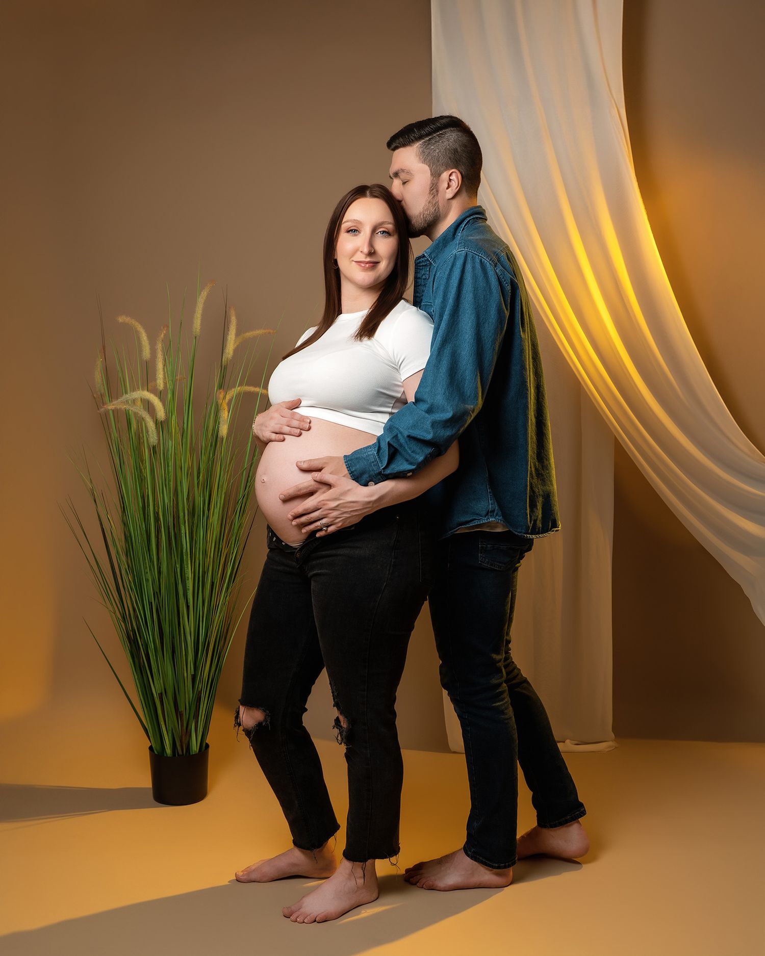 Modern maternity portrait with couple in casual wear standing against warm-toned studio backdrop.