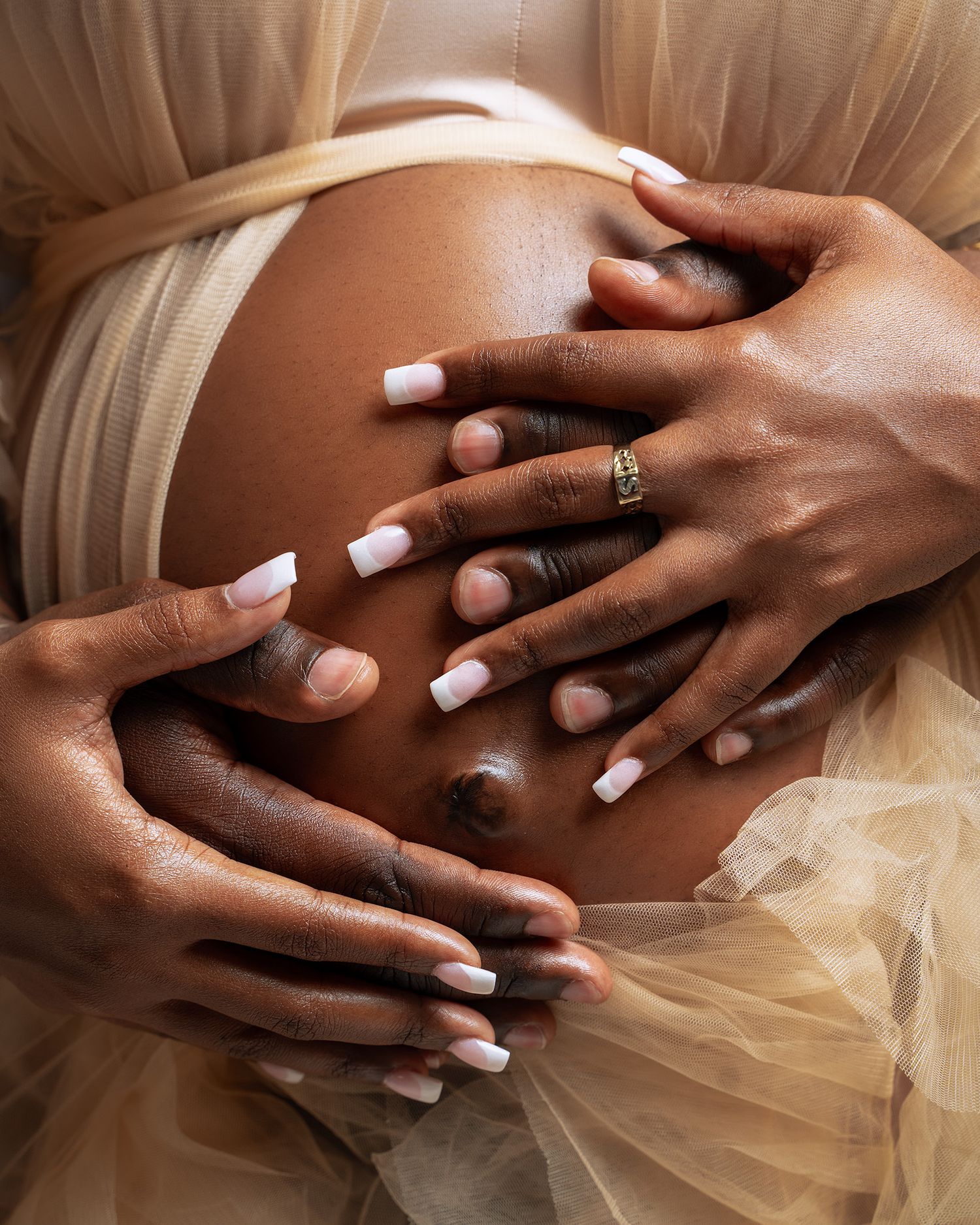 Close up of hands with manicured nails resting on a pregnant belly against tulle fabric.