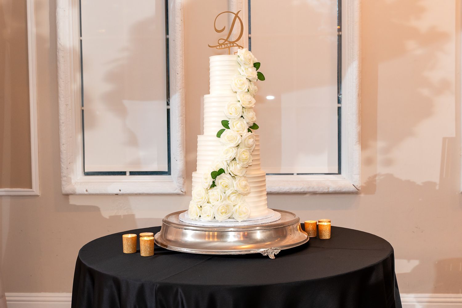 Elegant white wedding cake with monogram D and greenery on black tablecloth.