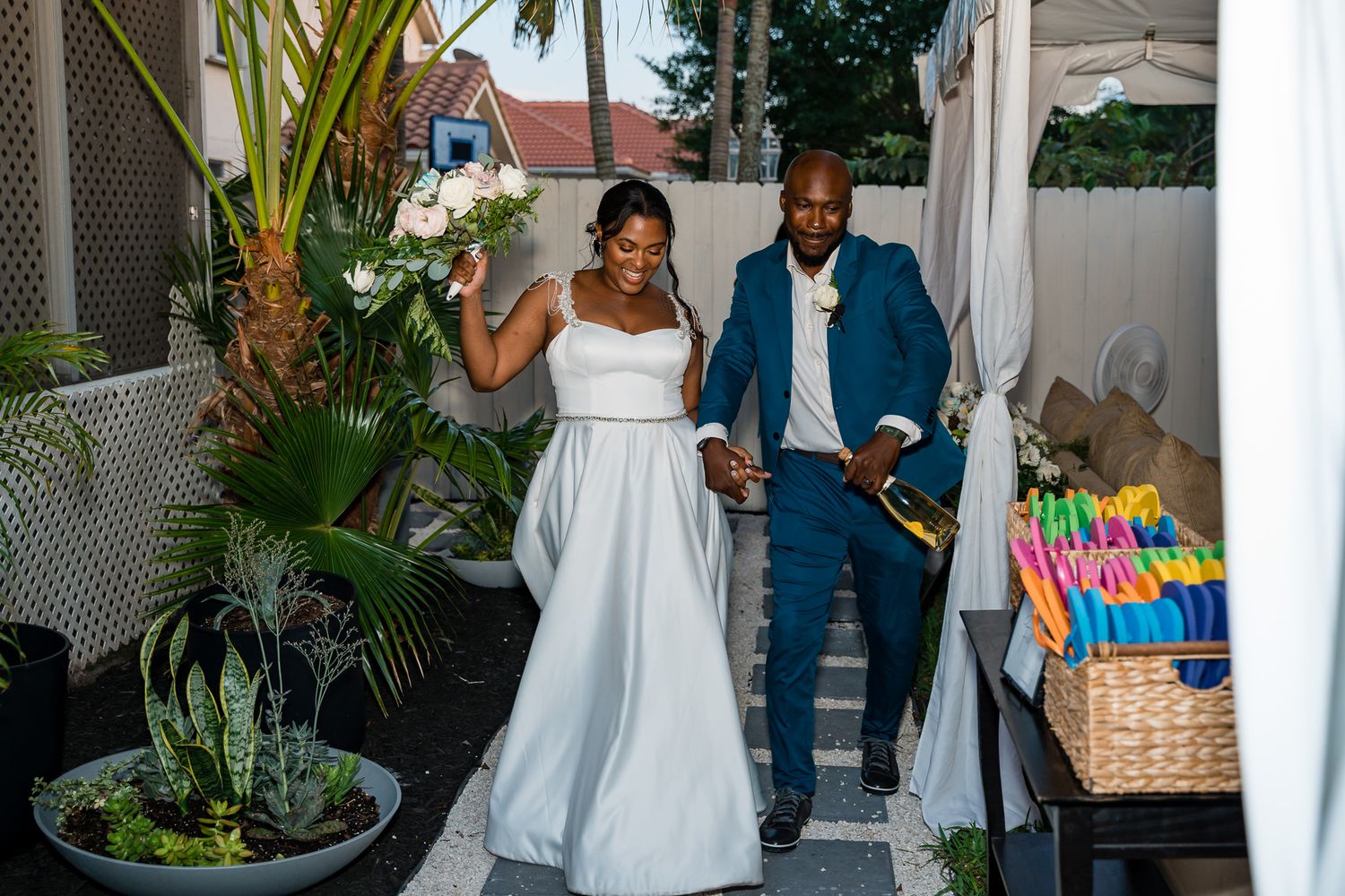 Bride in white dress and groom in blue suit walking through garden patio with tropical plants.