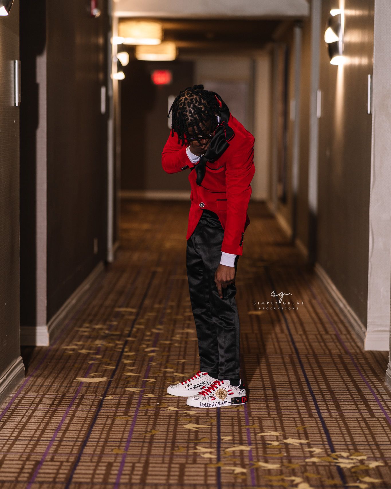 Person in red blazer and black pants looking down at designer sneakers in hotel hallway.