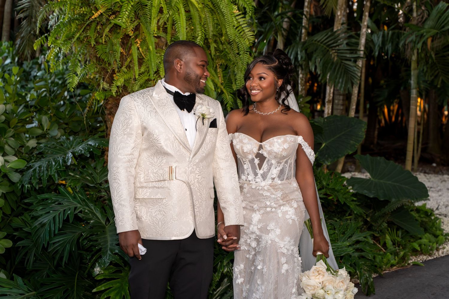 A couple poses together in an elegant outdoor garden setting wearing formal white wedding attire.