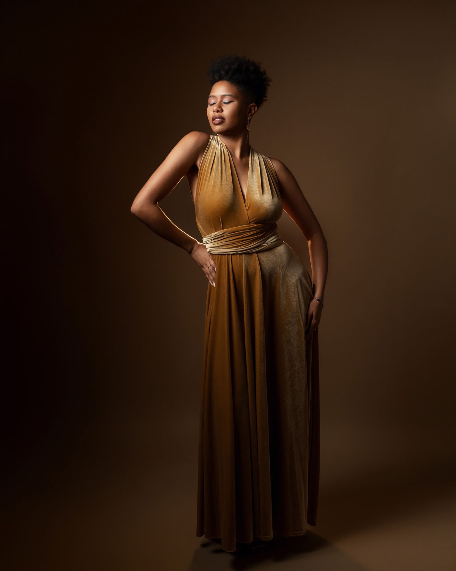Elegant full-length portrait in gold halter dress against brown background.