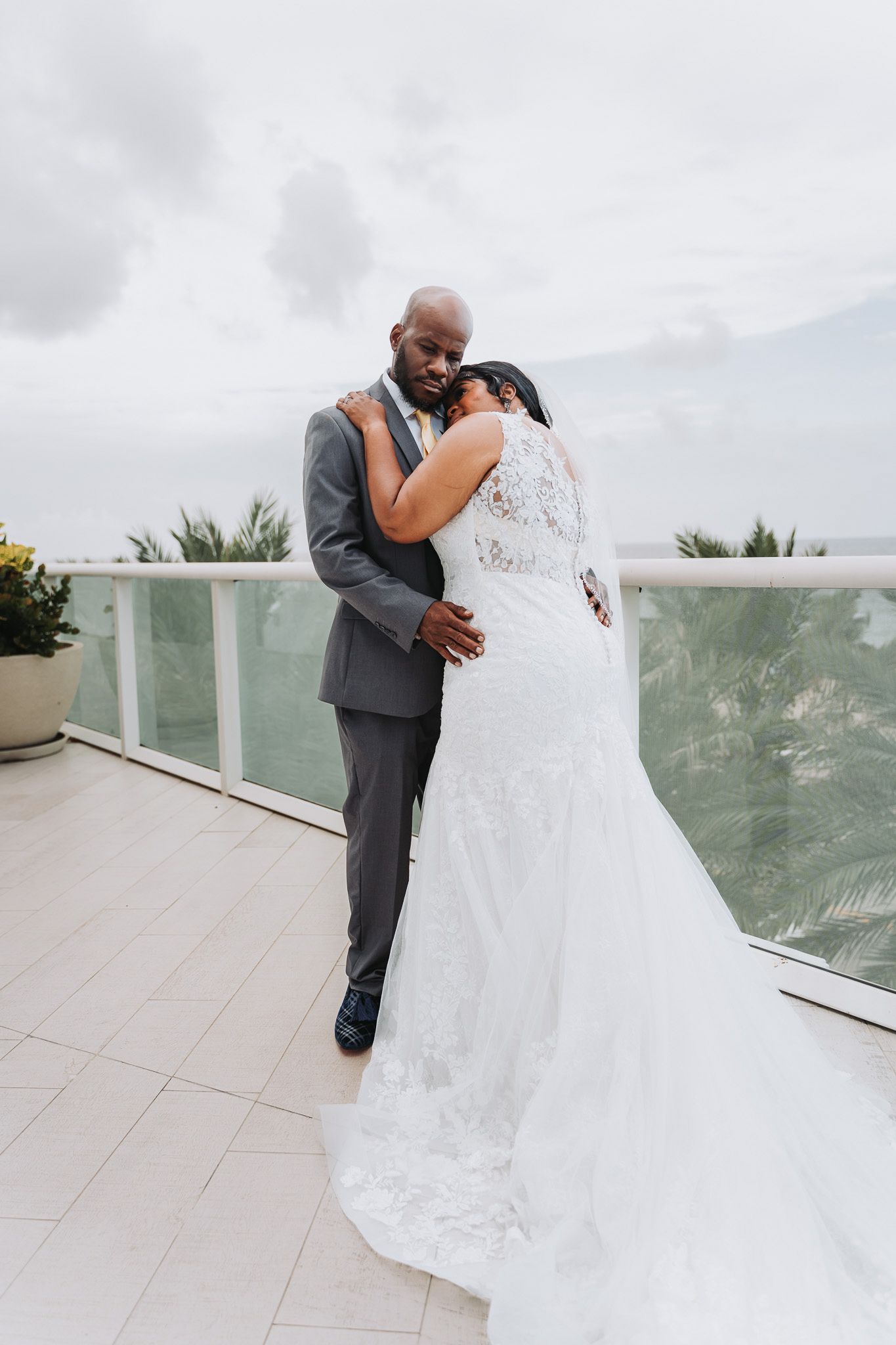 Newlywed couple embracing on a balcony overlooking palm trees, with bride in white lace gown and groom in grey suit.