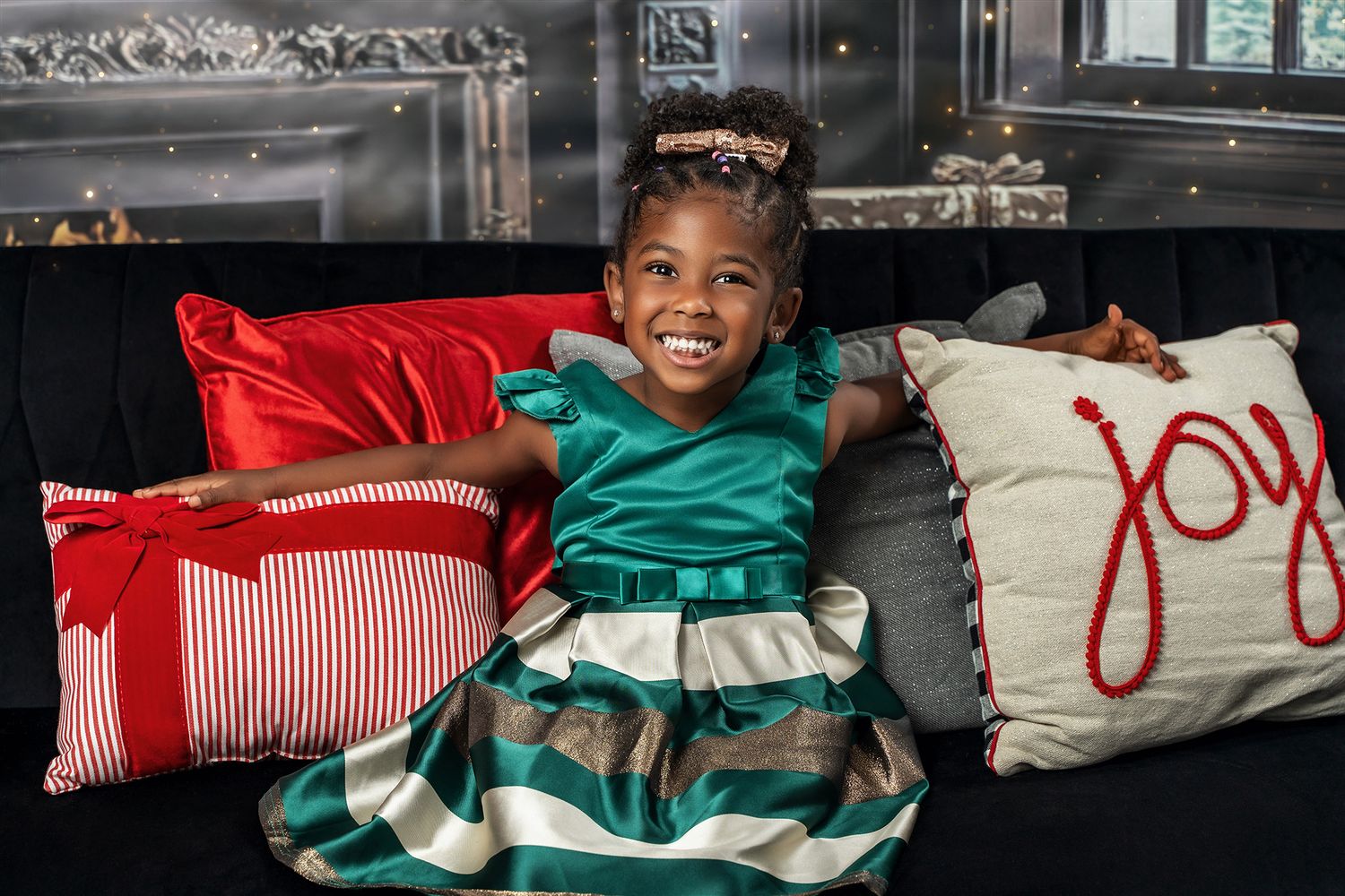 A child in a festive emerald green dress sits happily on a couch with decorative holiday pillows.