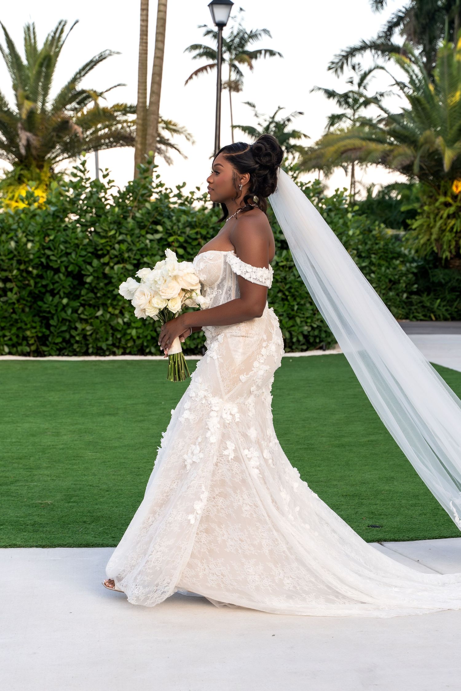 A bride in an elegant off-shoulder lace wedding gown holds a white bouquet against a tropical garden backdrop.