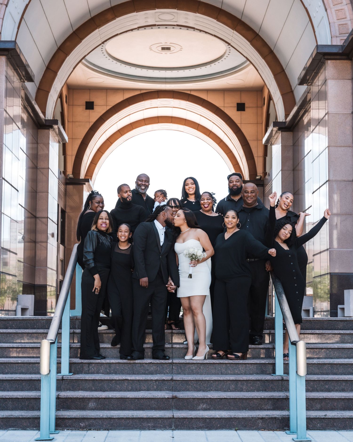 A celebratory group photo of a wedding party posing joyfully on elegant stone steps under arches.