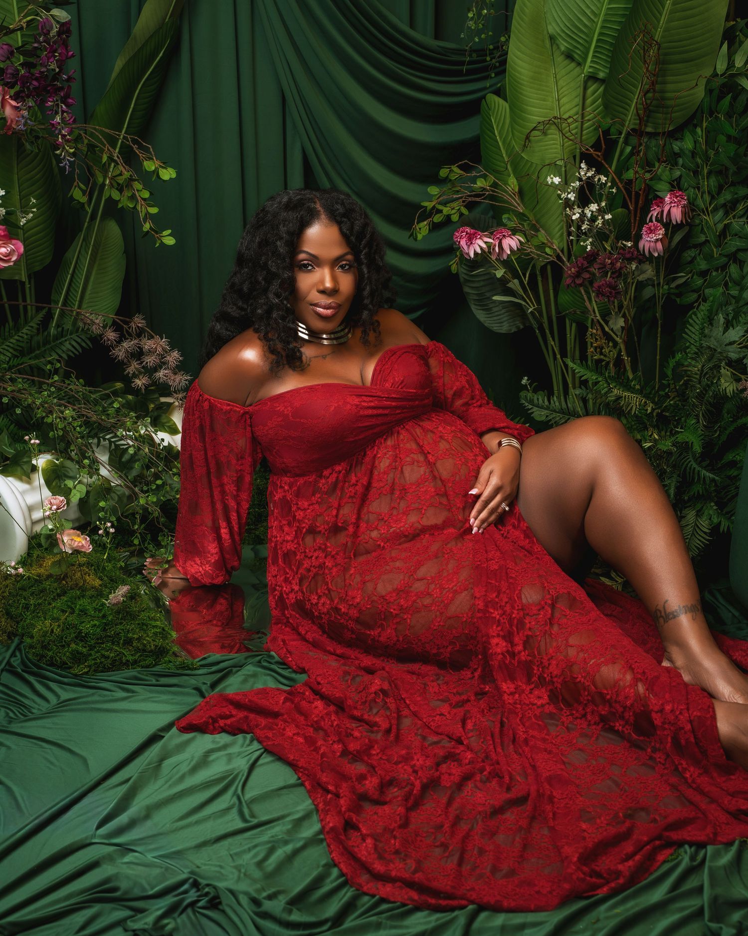 A figure in a flowing red lace dress poses elegantly against a lush tropical backdrop with green foliage and pink flowers.