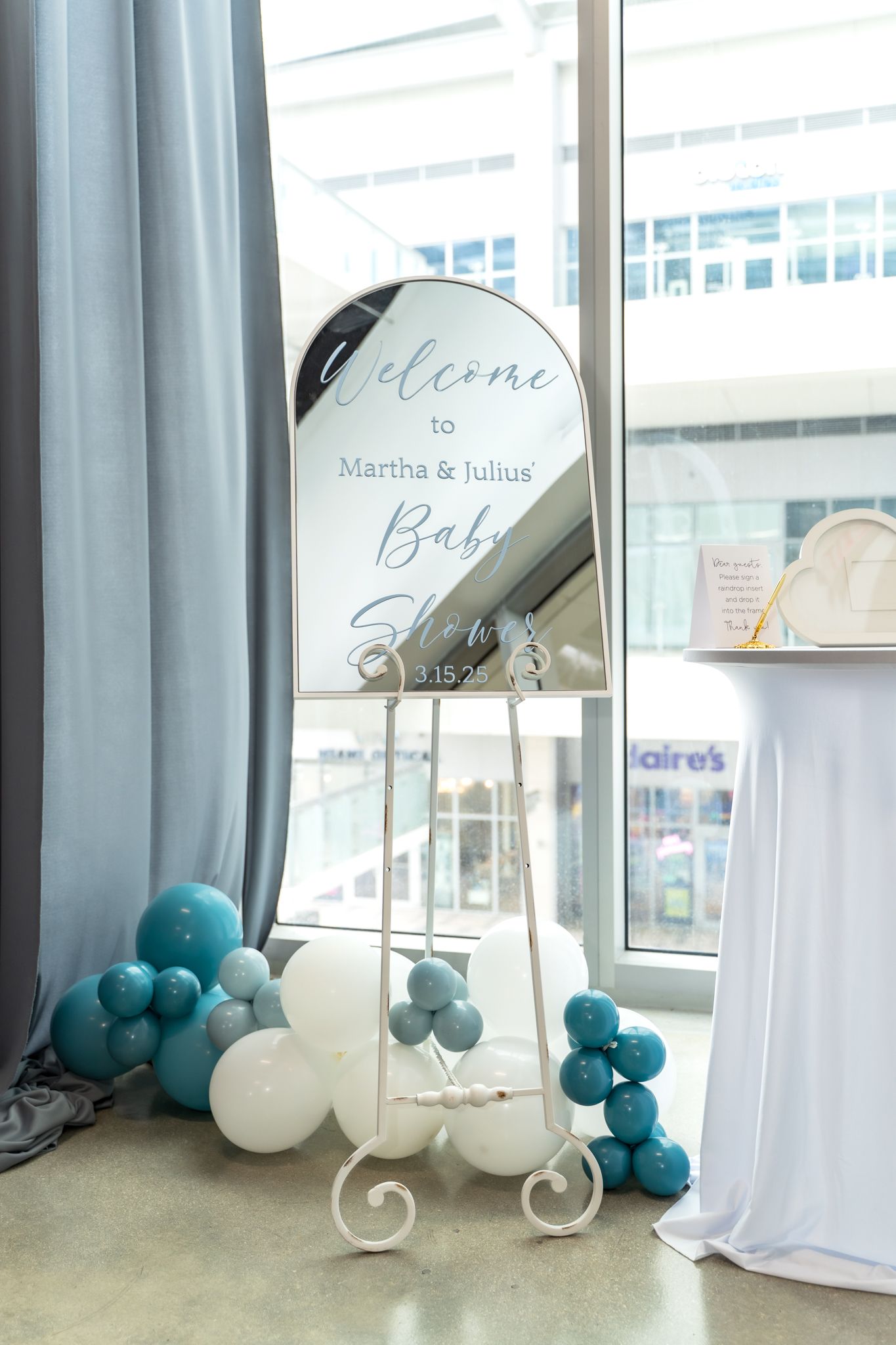 Welcome sign on decorative stand with blue and white balloon garland for baby shower.
