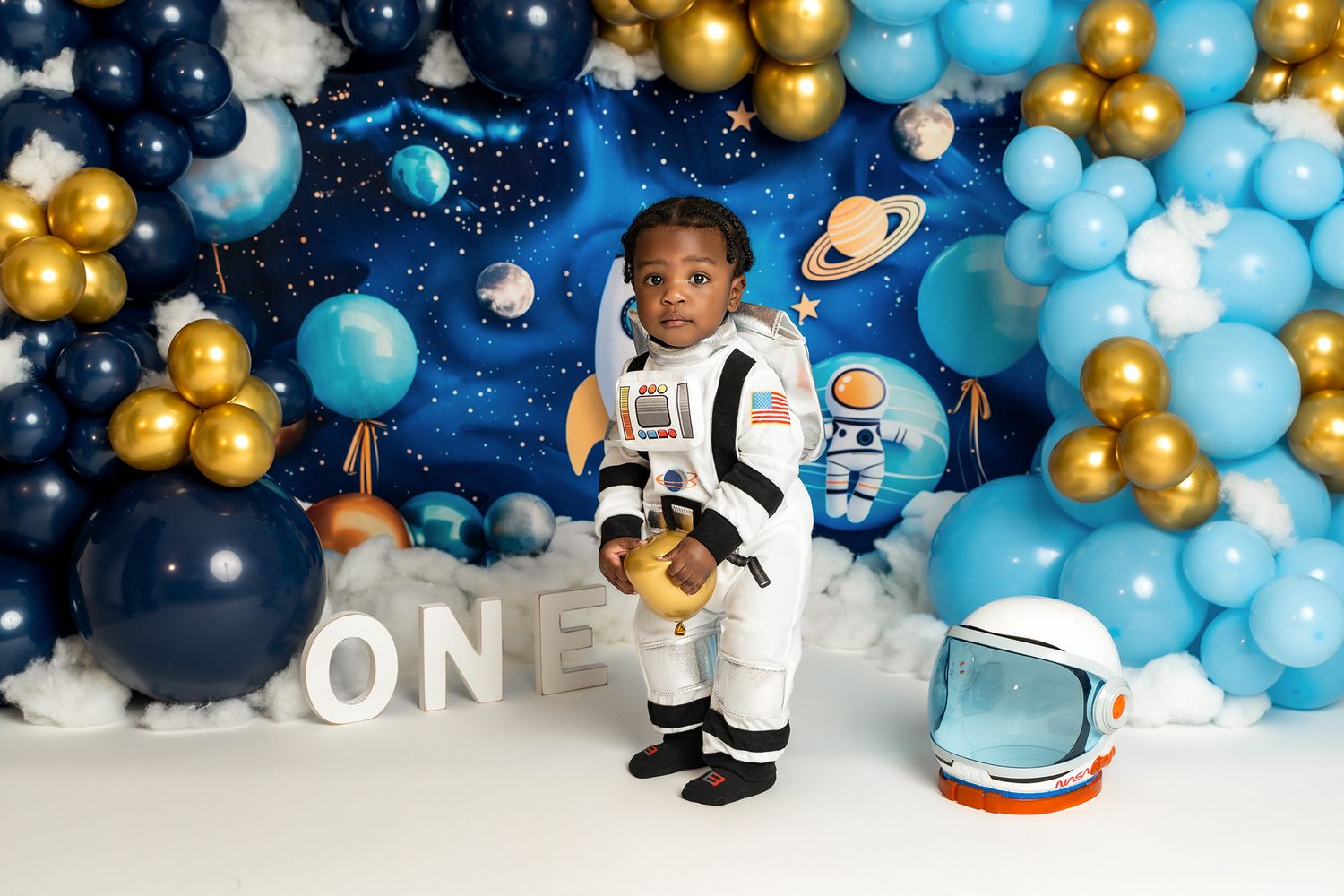 A toddler in a white space suit poses near a decorative helmet against a galaxy-themed balloon backdrop.