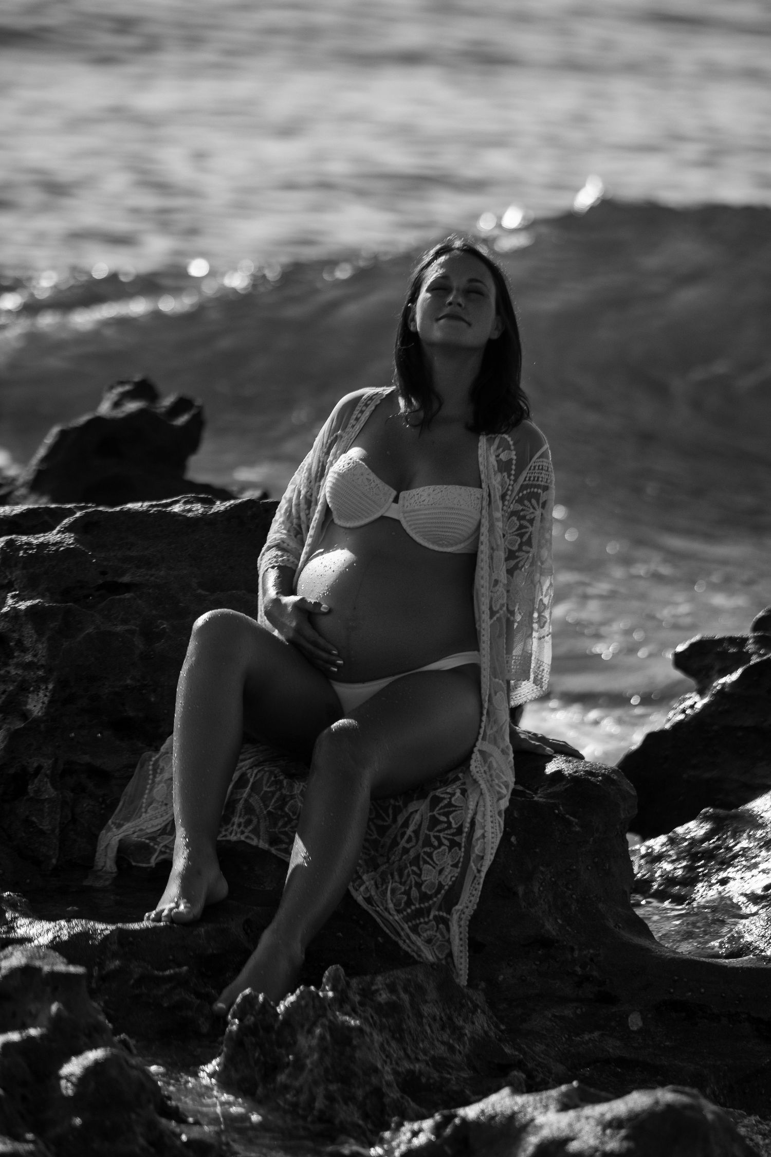 Artistic black and white maternity photo of an expectant mother in white swimwear and lace robe on coastal rocks.