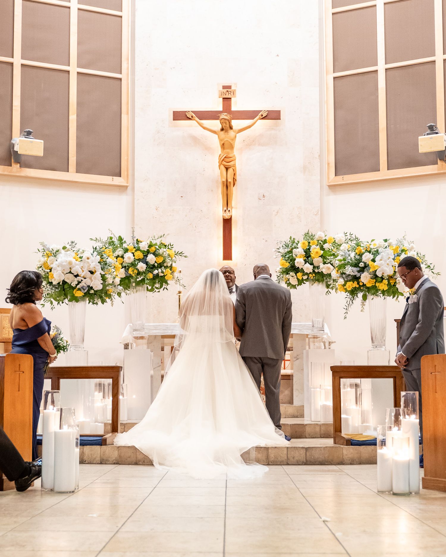 Wedding ceremony in church with crucifix and floral arrangements at altar.