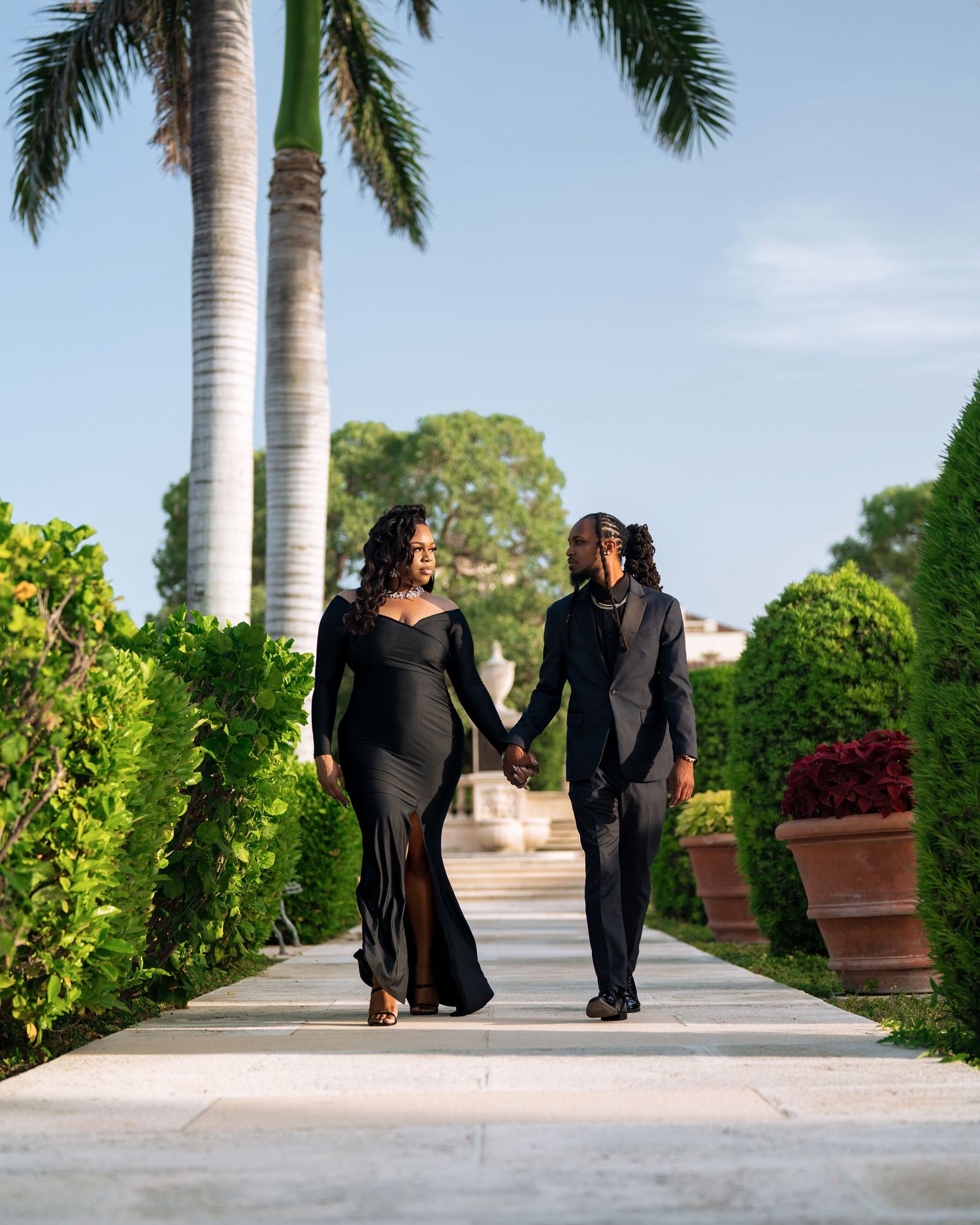 Three sequential views show figures strolling along a palm-lined pathway in formal black attire at a tropical venue.