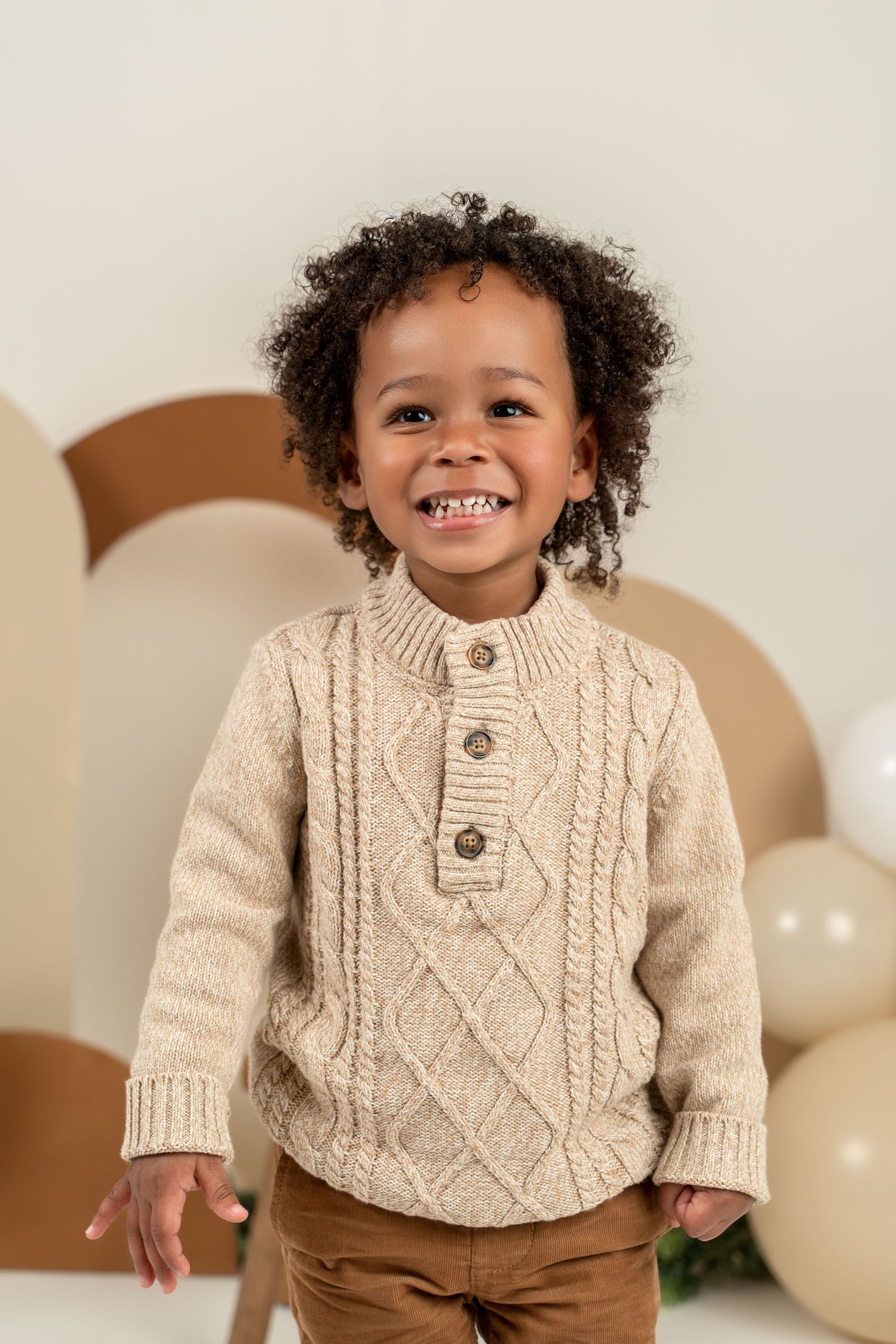 Smiling child wearing a cozy cream cable knit sweater poses against neutral background with balloons.