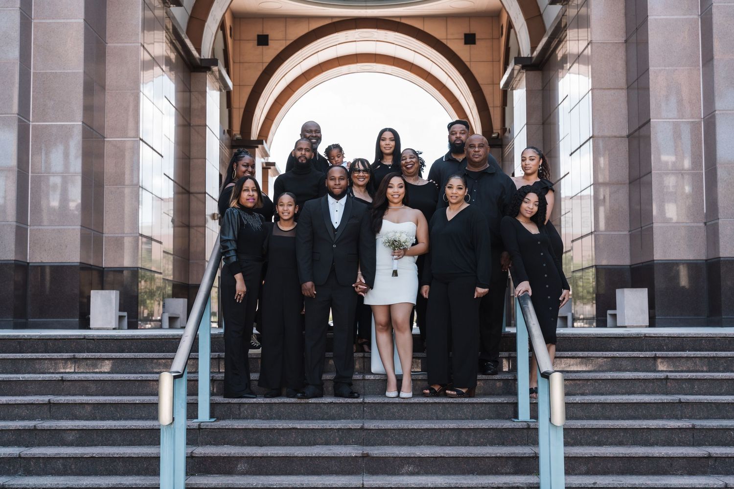 A wedding party in black attire and one person in white standing on stone steps under an arched entryway.
