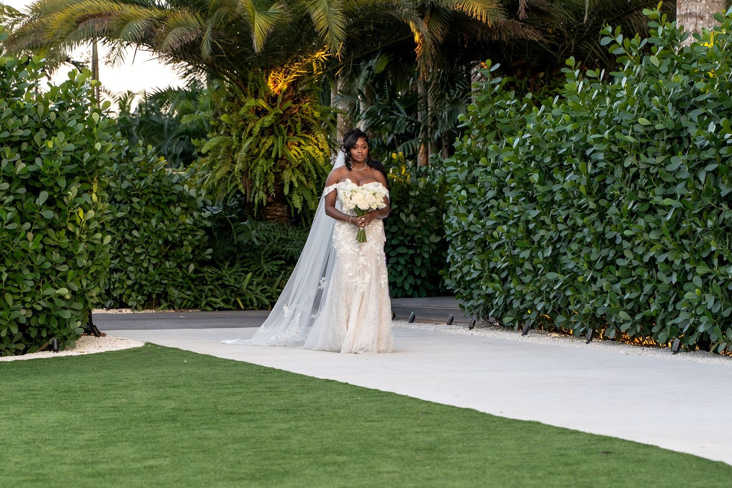 A person in a white wedding dress poses in a lush tropical garden setting with palm trees and greenery.