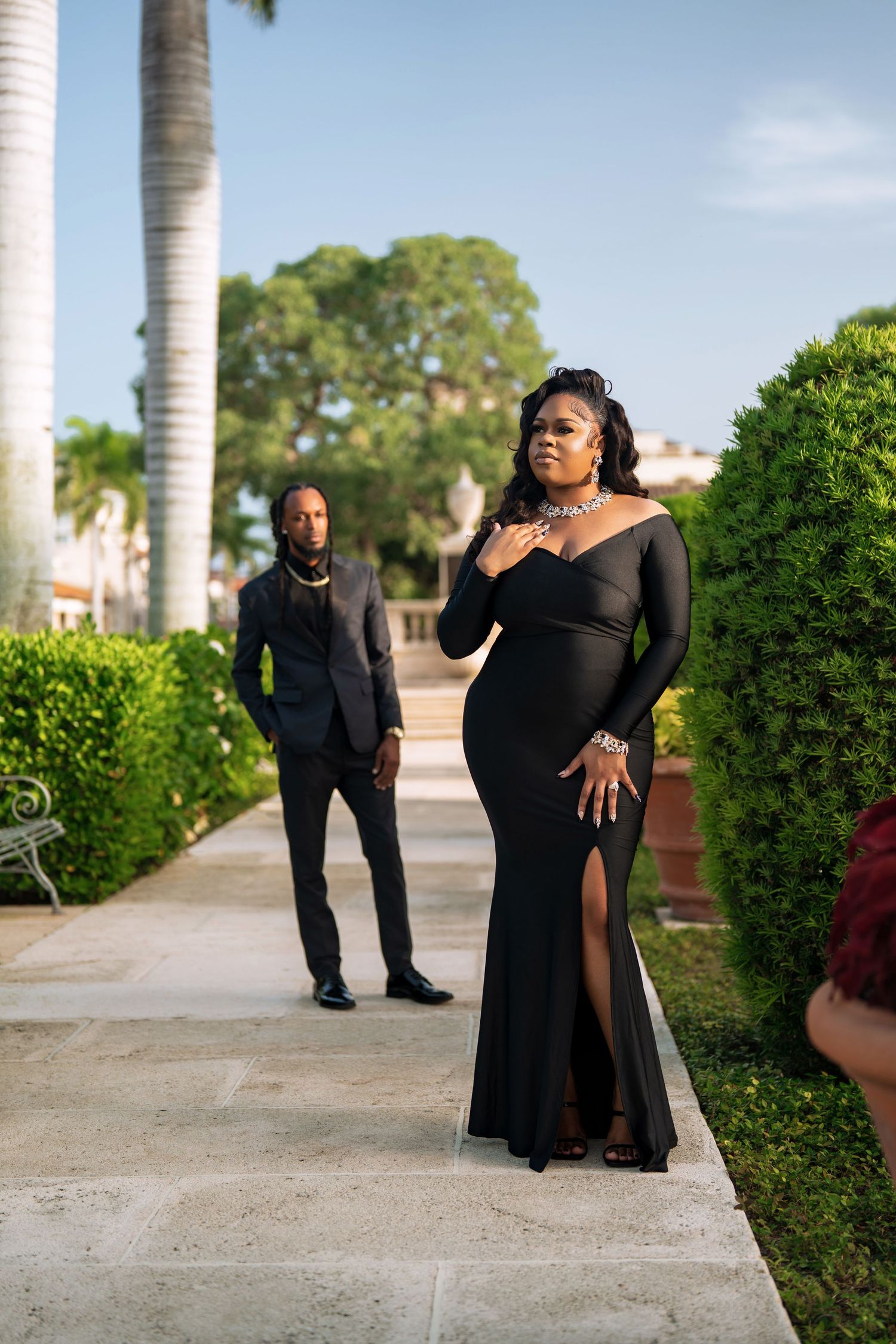 A couple in elegant black formal attire poses on a palm-lined walkway for a glamorous outdoor photoshoot.