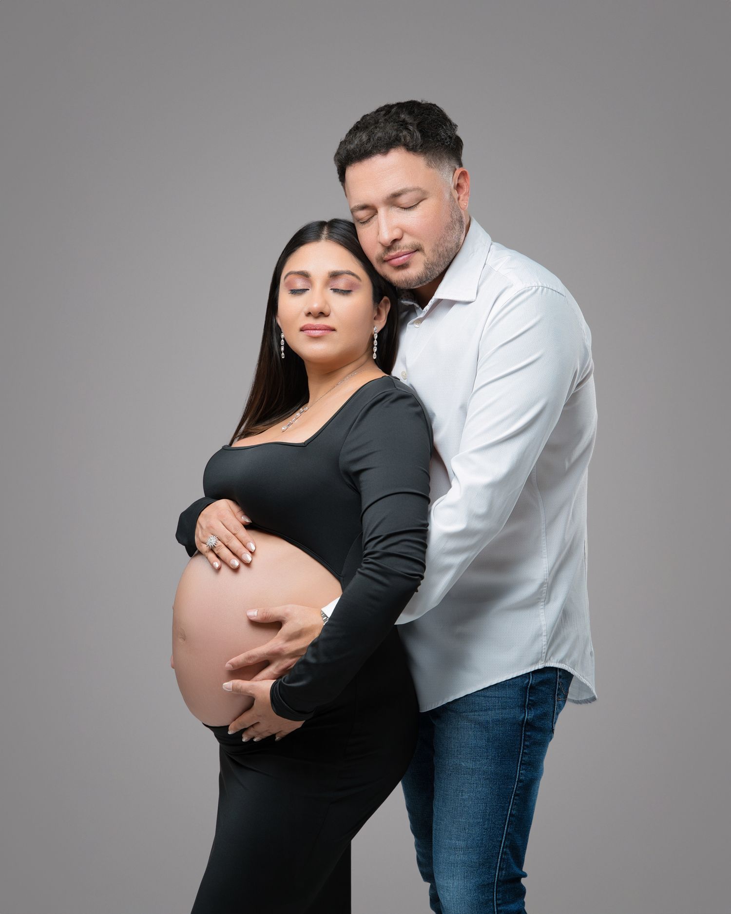 A maternity portrait against grey background shows an expectant couple in a tender embrace.