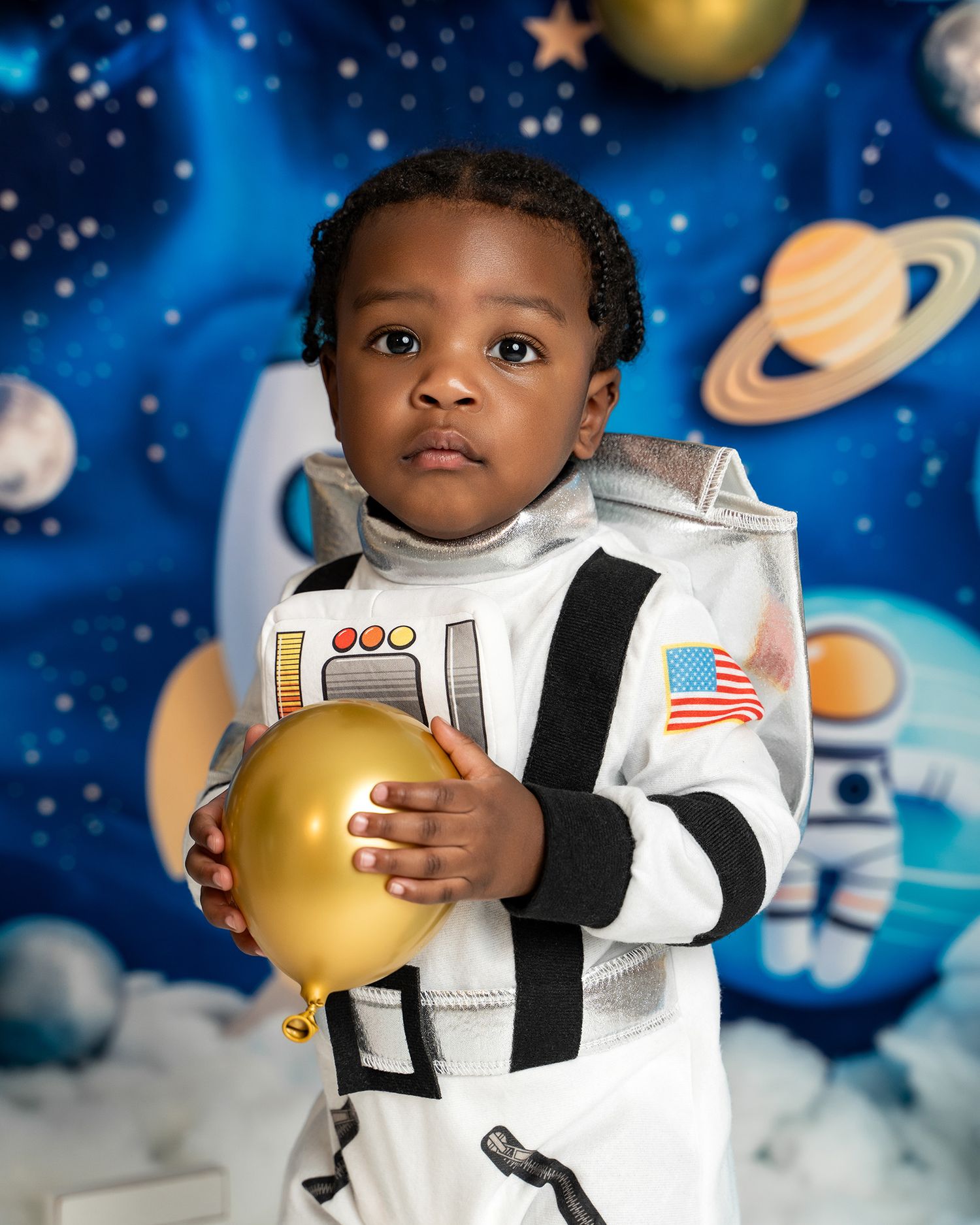 A close-up portrait of a child in an astronaut costume holding a gold balloon against a starry backdrop.