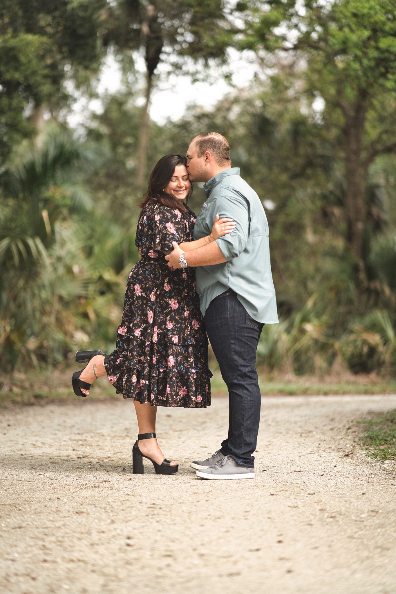 Romantic couple embracing on a nature path, with her floral dress and his blue shirt creating a sweet moment.