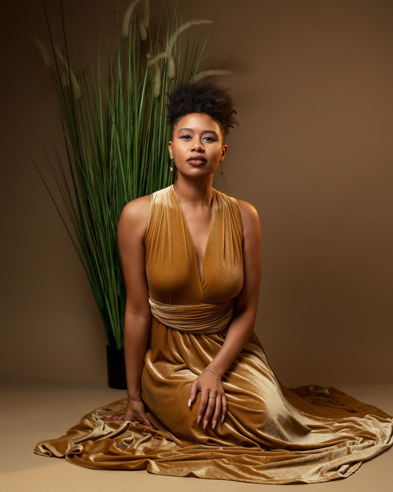 Sophisticated portrait with decorative plants wearing gold evening gown against earthtone background.