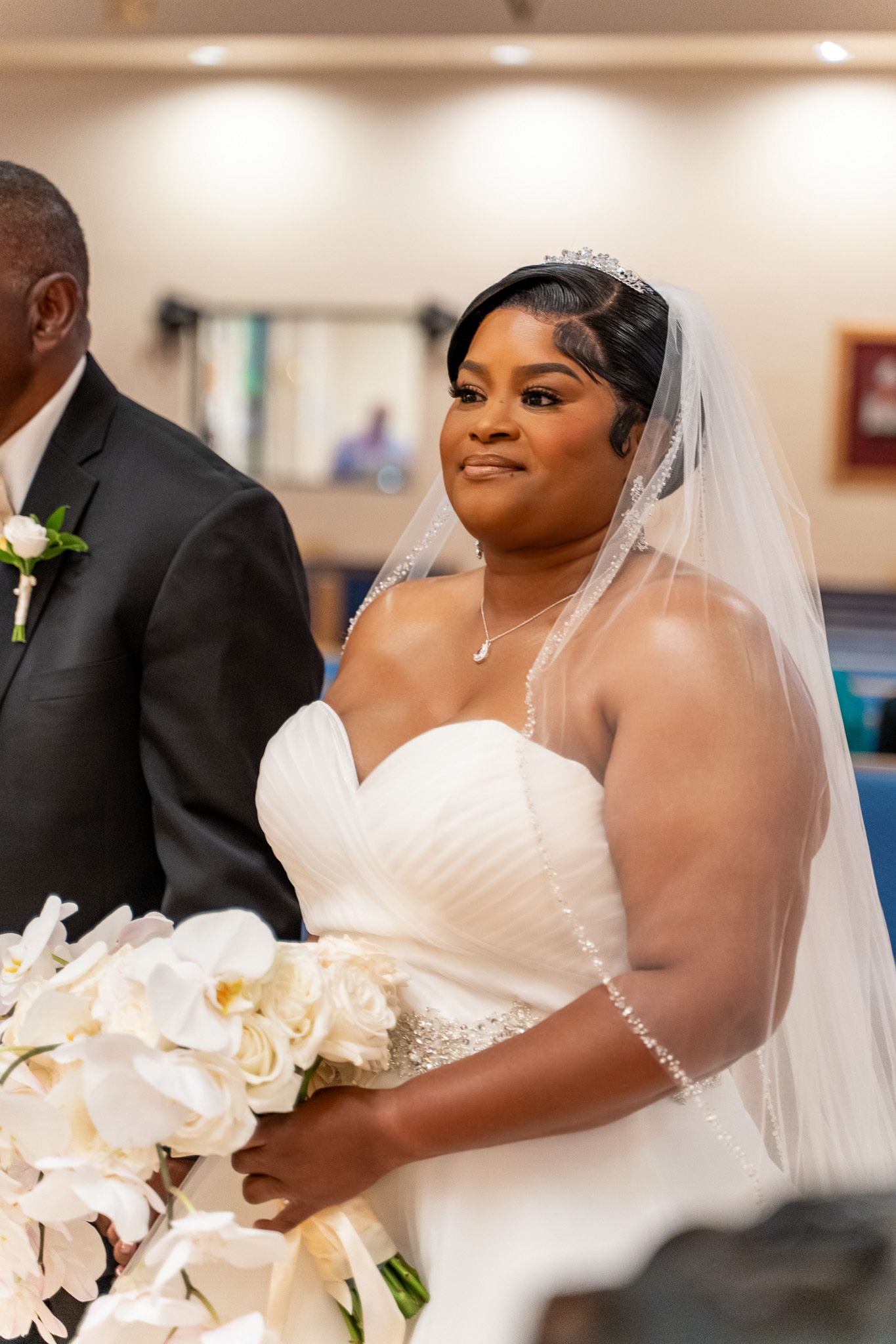 Bride wearing veil and strapless wedding dress stands with bouquet during ceremony.