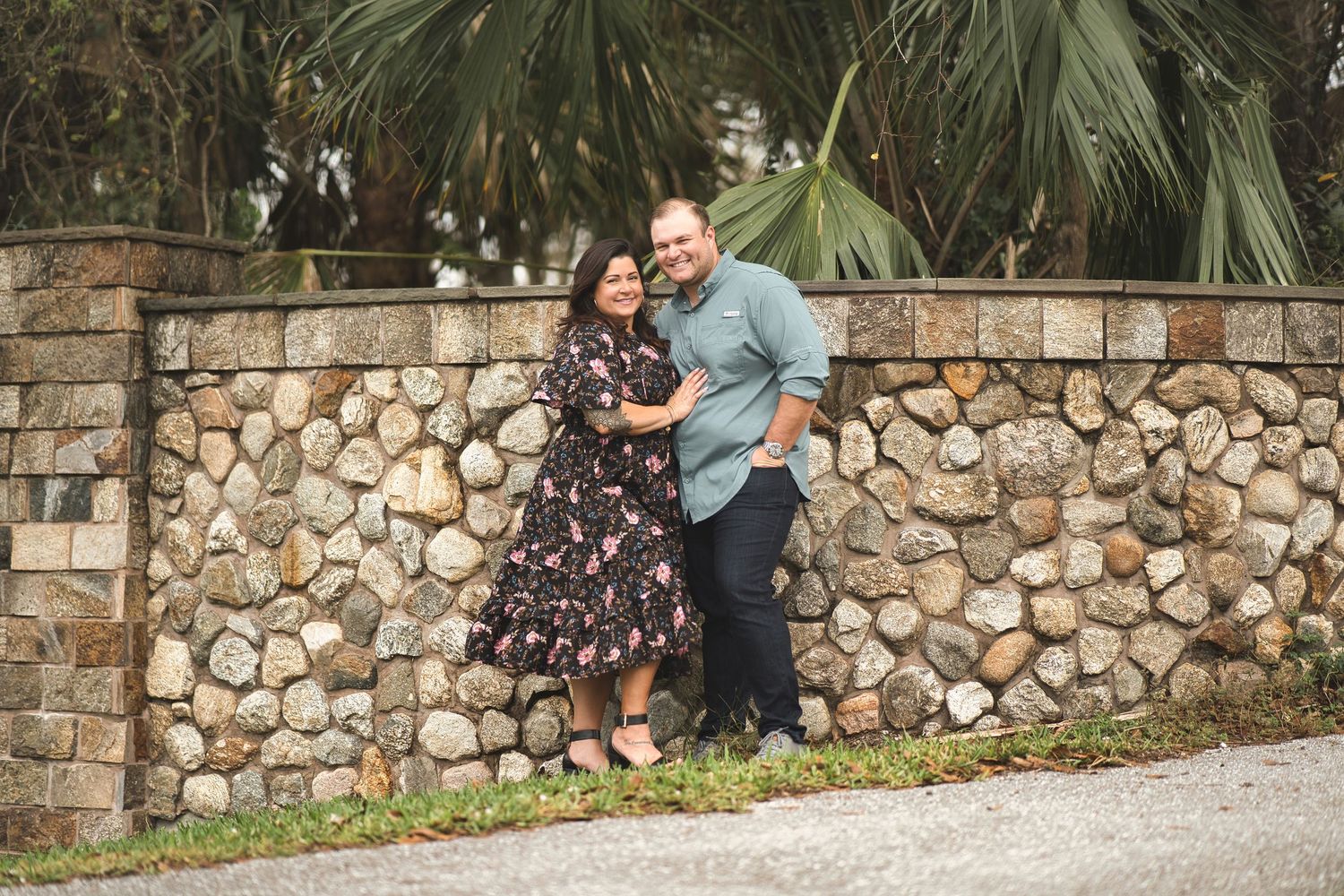 Couple posing against a rustic stone wall with palm trees in background.
