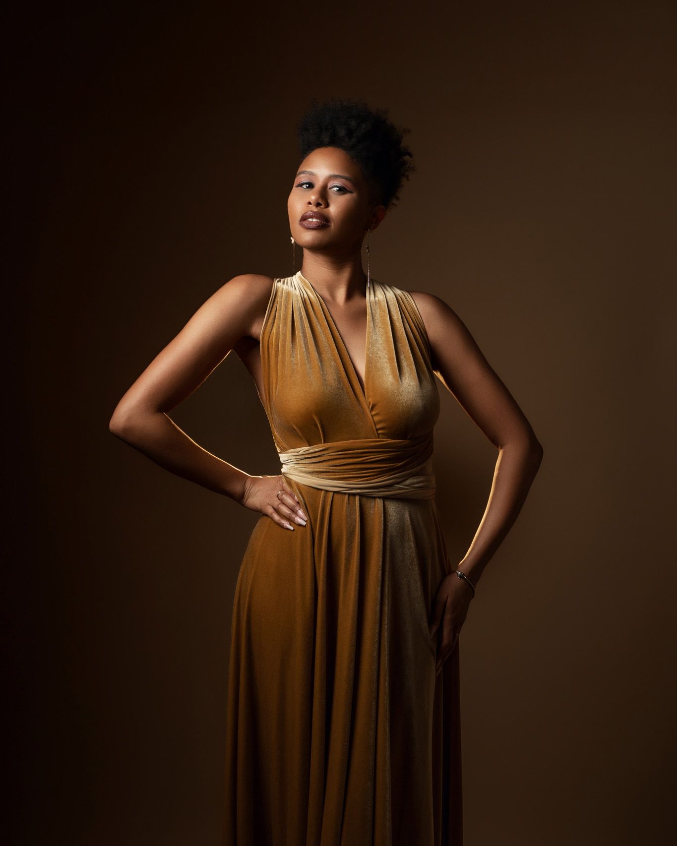 Elegant studio portrait of woman in flowing gold wrap dress against brown backdrop.