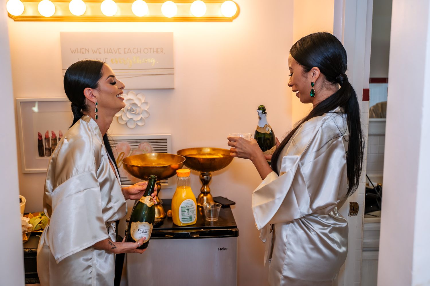 Two people in white satin robes getting ready in a dressing room with vanity lights.