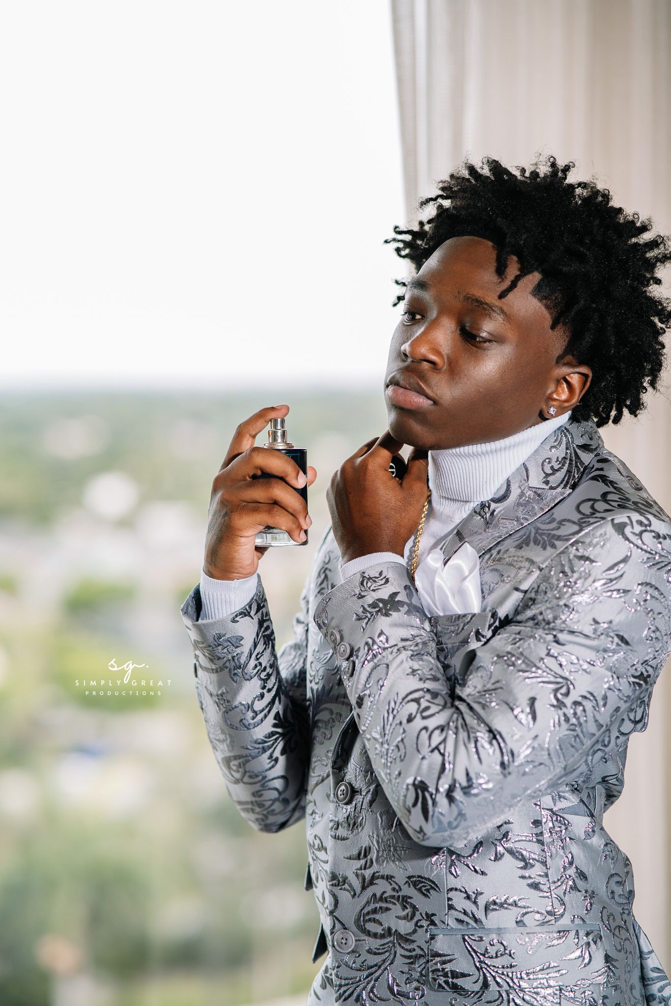 Person wearing silver patterned suit jacket and white turtleneck poses by window.