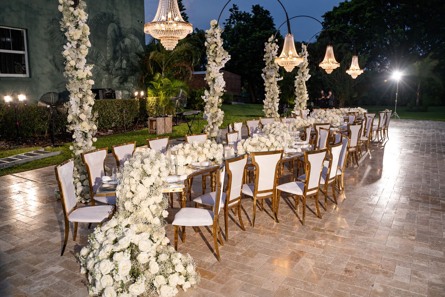 An outdoor wedding setup at dusk with white floral arches and cross-back chairs illuminated by string lights.