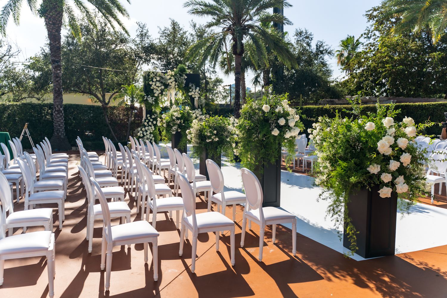White chairs arranged in rows for an outdoor wedding ceremony with palm trees and tropical landscaping.