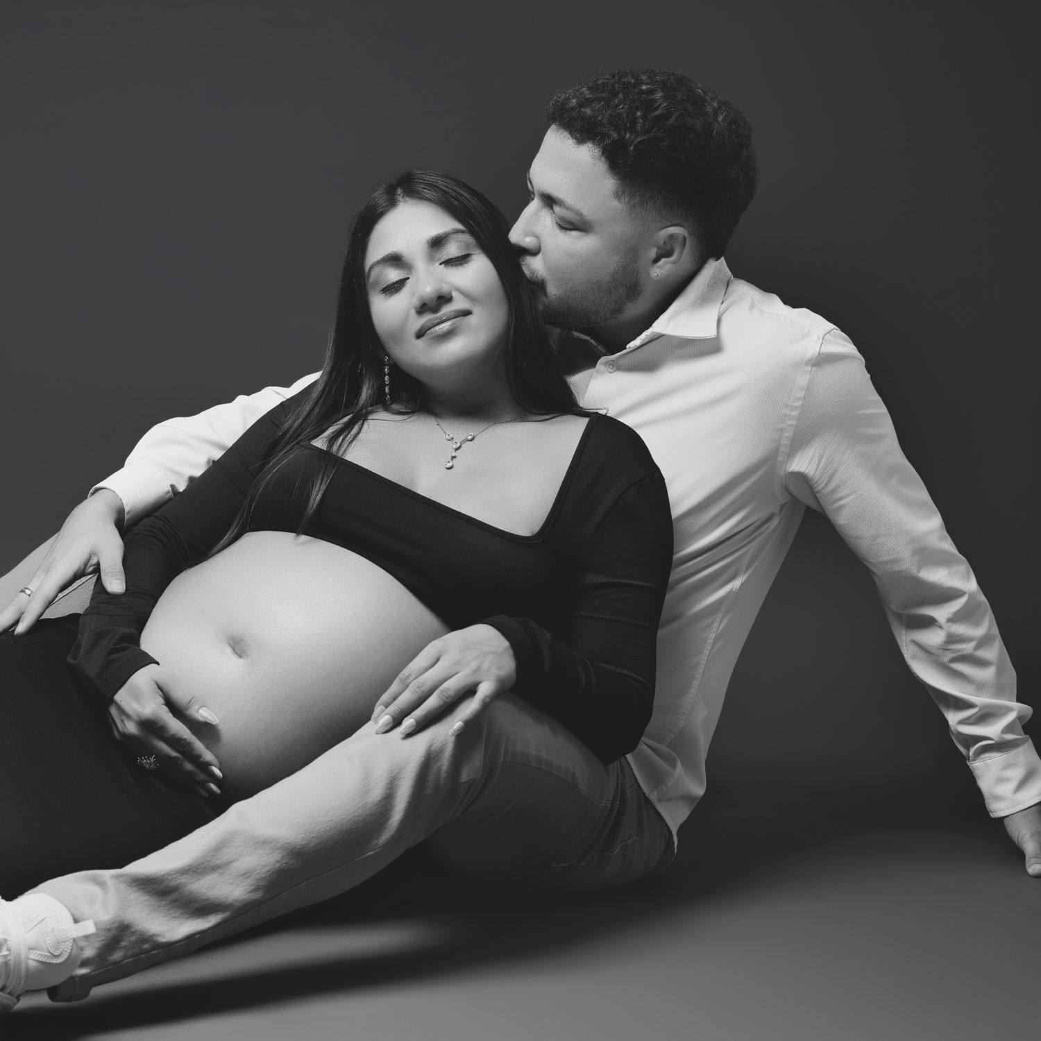 Black and white maternity portrait of couple in intimate pose on dark background.
