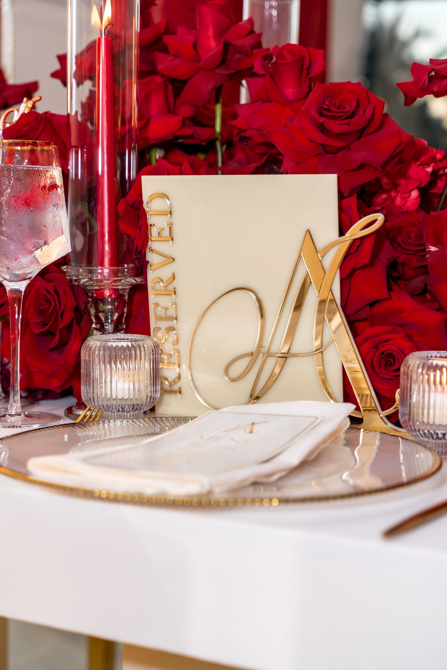 Elegant wedding table setting with red roses and gold accents featuring place cards and crystal glassware.