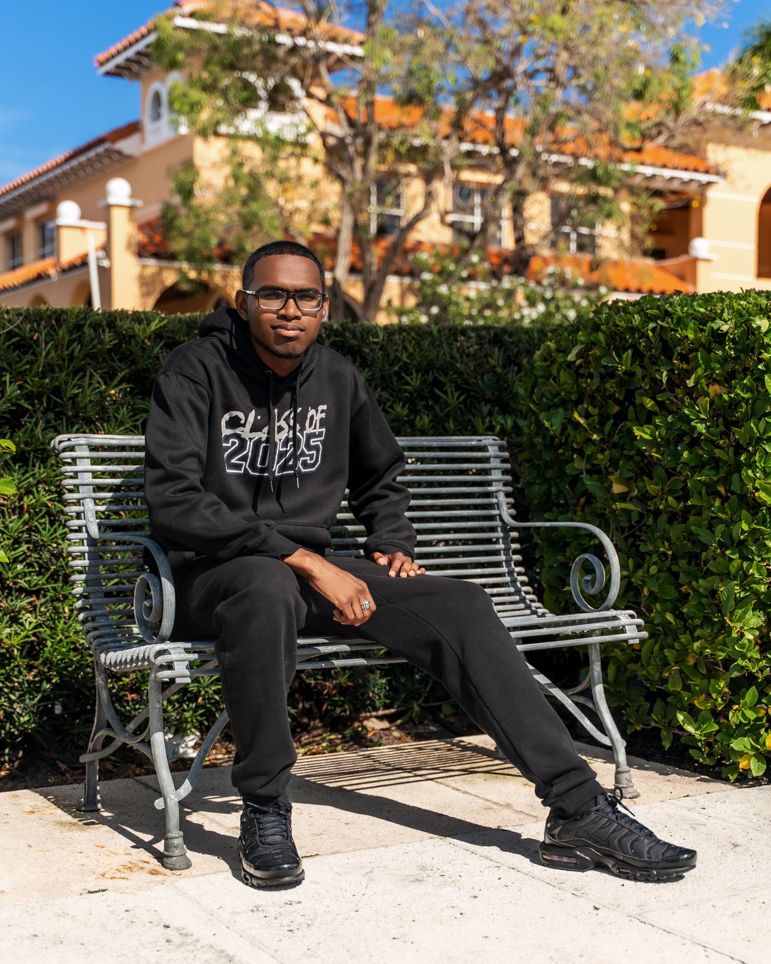 Someone in a black Chase 2023 hoodie and pants sits relaxed on a metal bench in front of Spanish-style architecture.