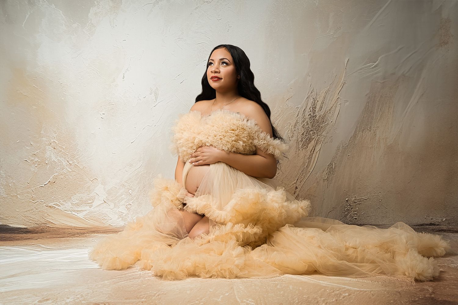Elegant maternity portrait with figure seated in beige tulle gown against neutral background.