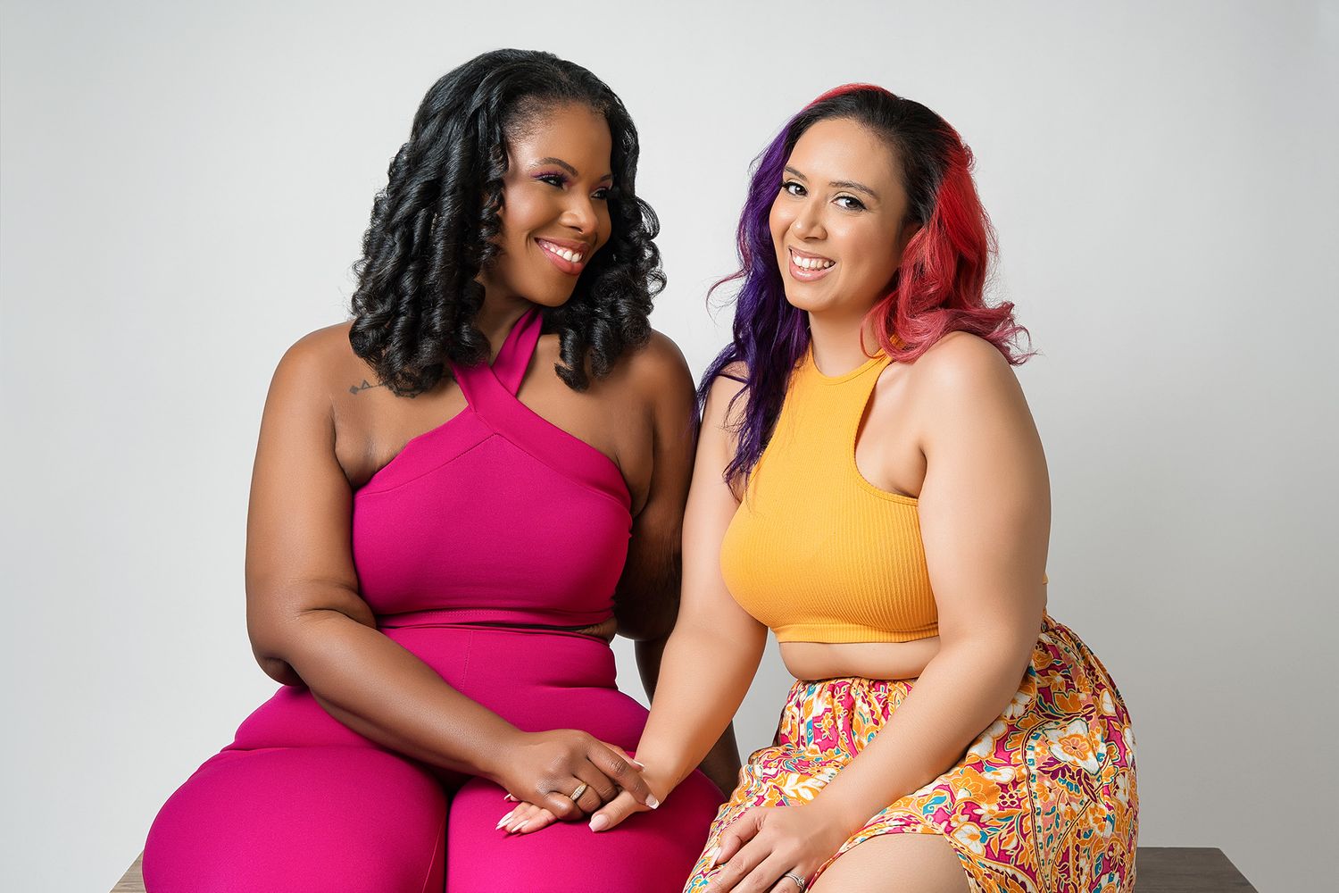 Models in vibrant pink and yellow outfits share a joyful moment during studio photoshoot.