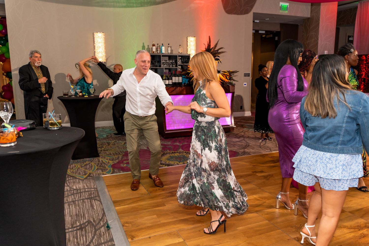 Party guests dancing together on a wooden dance floor with colorful mood lighting in the background.