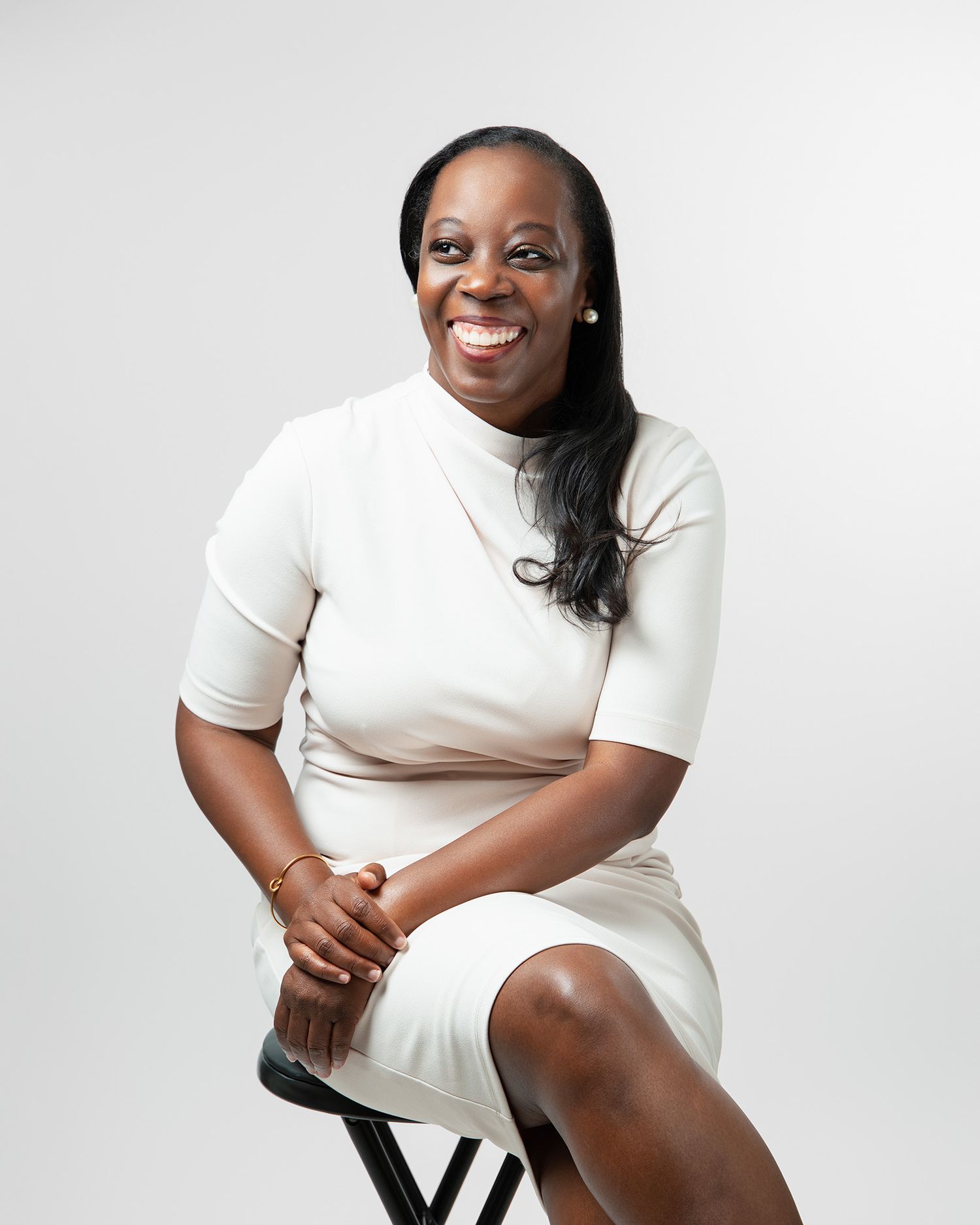 Professional portrait of someone in cream turtleneck dress seated on black stool.