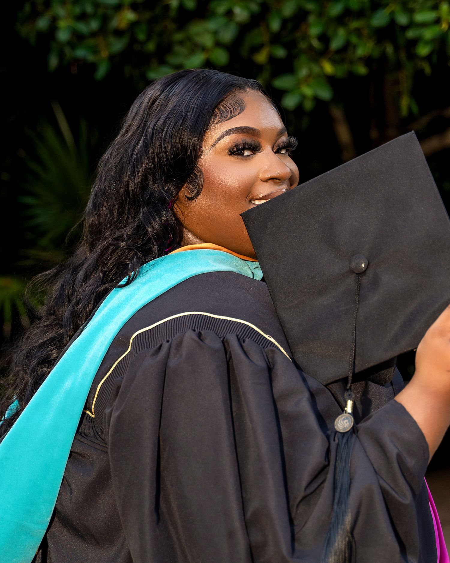 Graduate wearing academic cap and gown with teal stole smiles during graduation celebration.