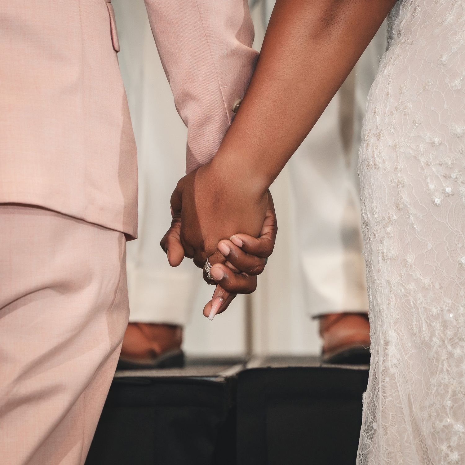Close-up shot of a couple holding hands with the groom in pink suit and bride in white lace dress.