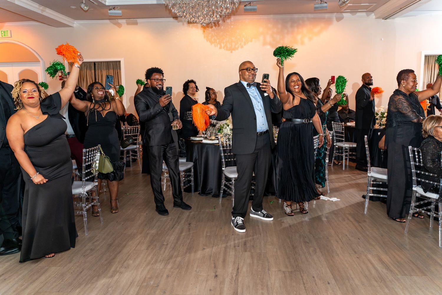 Wedding guests in formal black attire gather in an elegant ballroom with crystal chandeliers and hardwood floors.