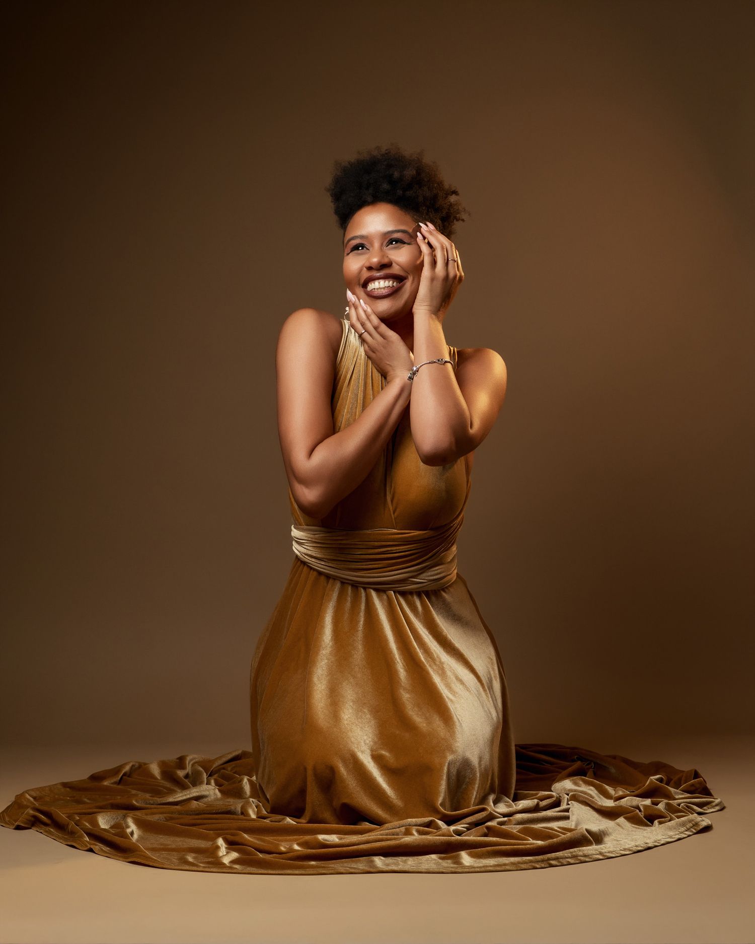 Fashion model in flowing gold satin dress smiles with hands on face against brown backdrop.