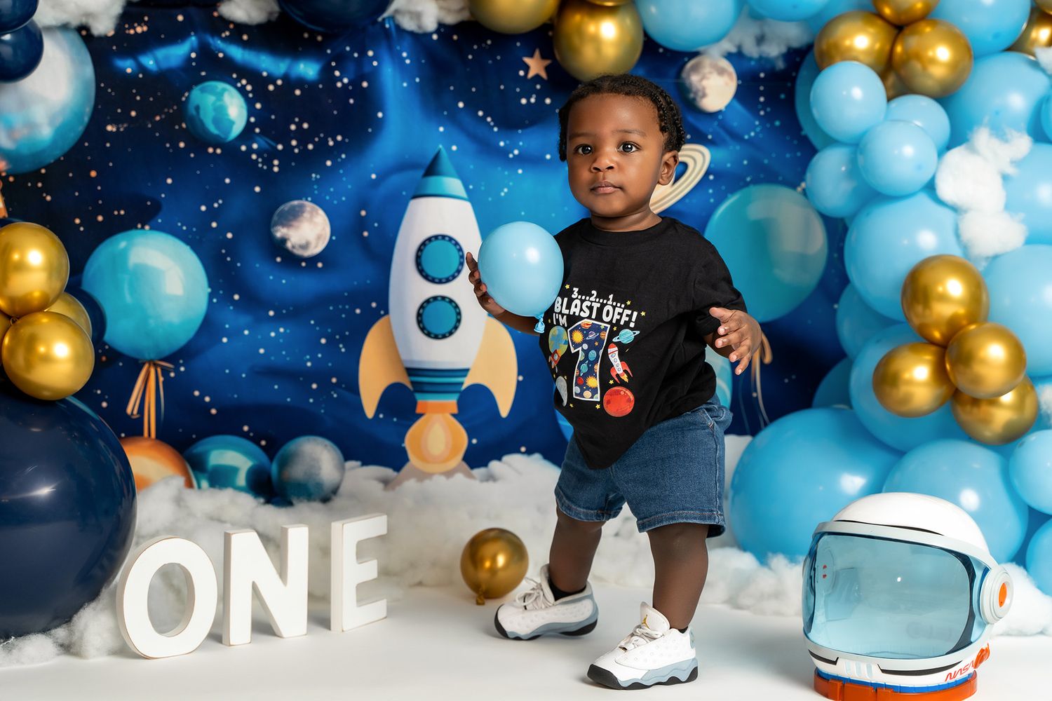 A toddler in a space-themed outfit stands with rocket ship decorations and colorful balloons for first birthday photos.