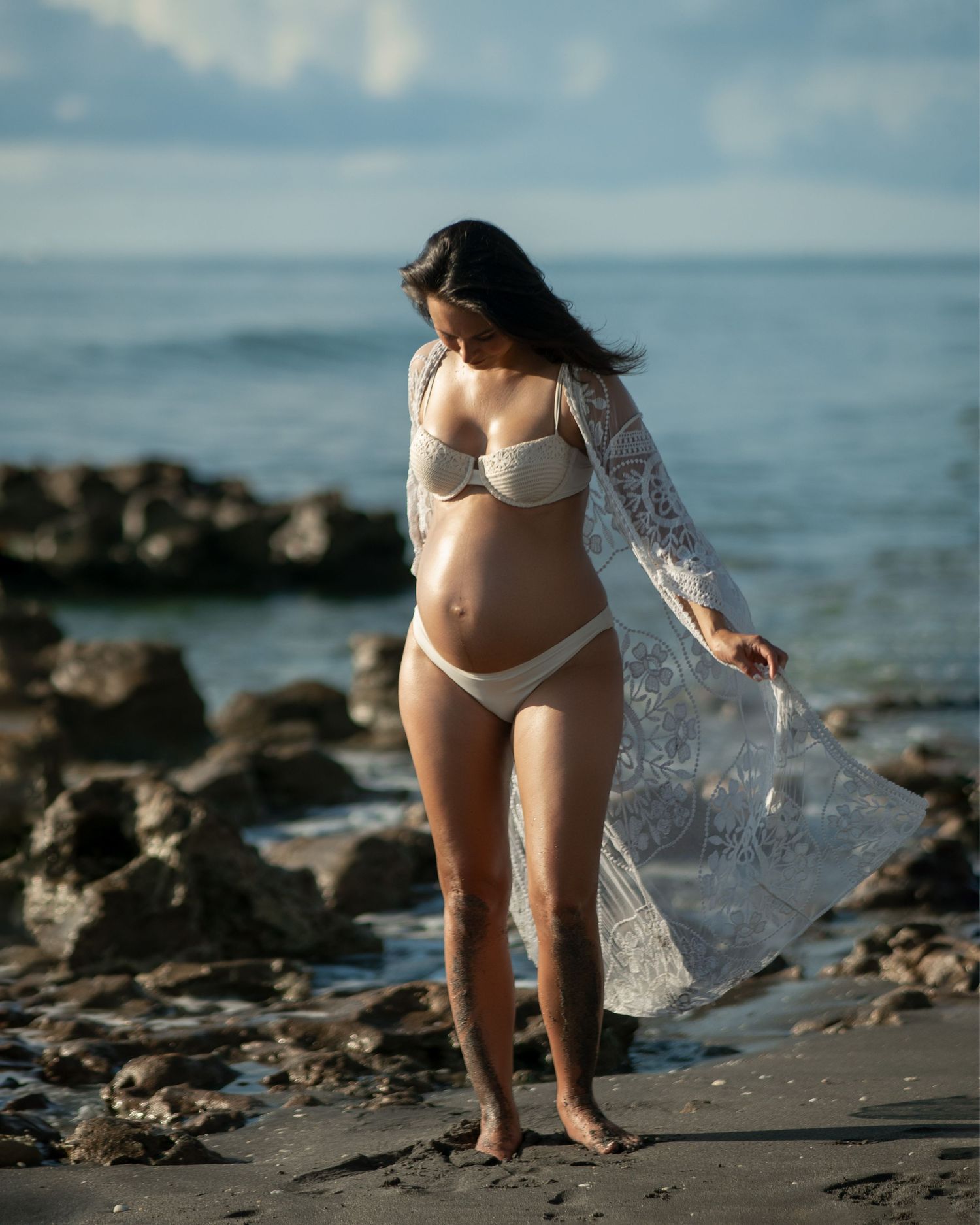 Elegant maternity beach photo with flowing lace robe catching ocean breeze.