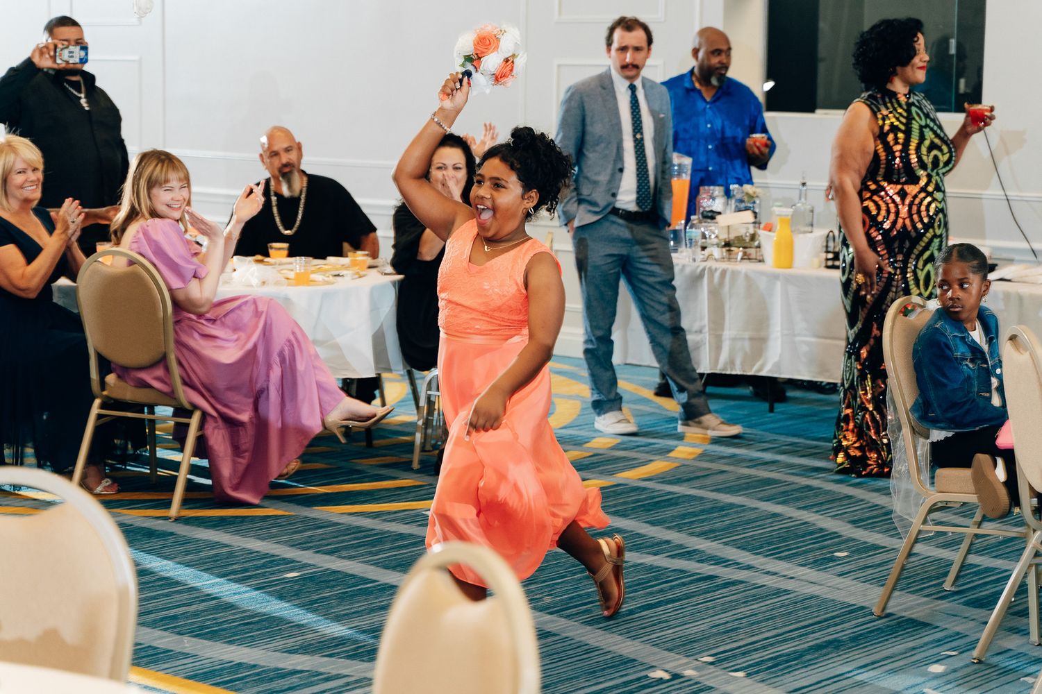 Guest in coral dress dances energetically at wedding reception with seated attendees in background.
