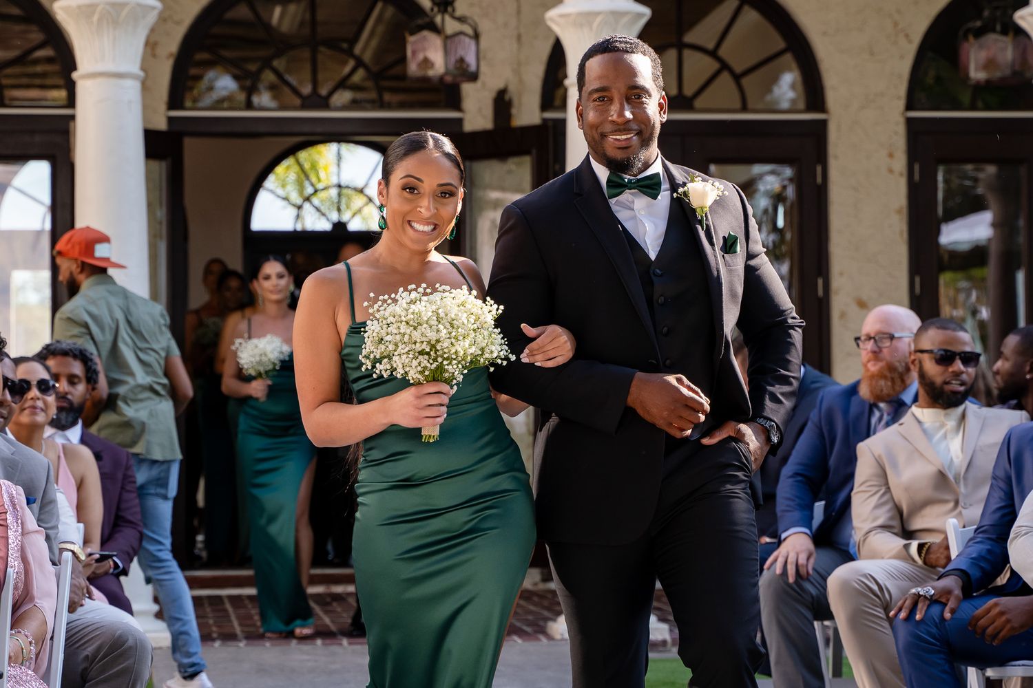 Formal wedding attendants walking down aisle in emerald green dress and black tuxedo.