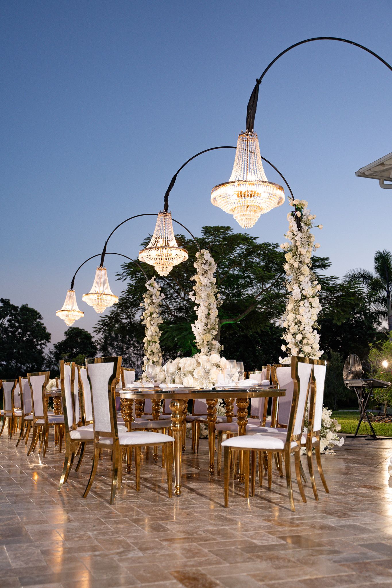 Luxurious outdoor wedding setup with crystal chandeliers and white floral arrangements on gold chairs.