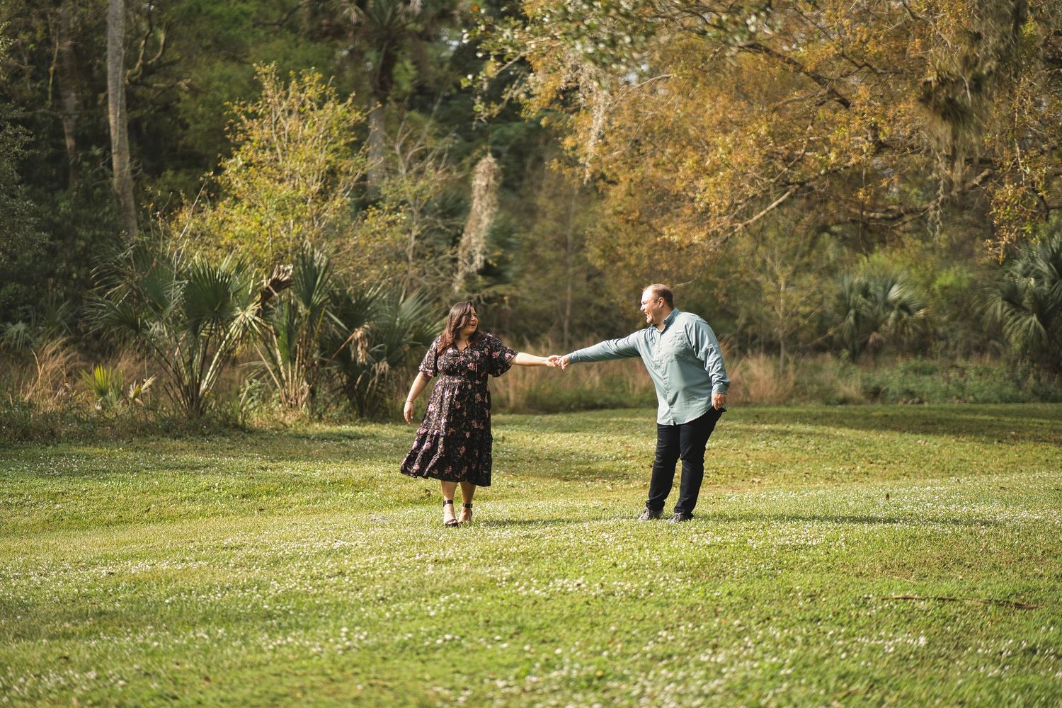 A couple dances playfully on a grassy field surrounded by autumn trees.